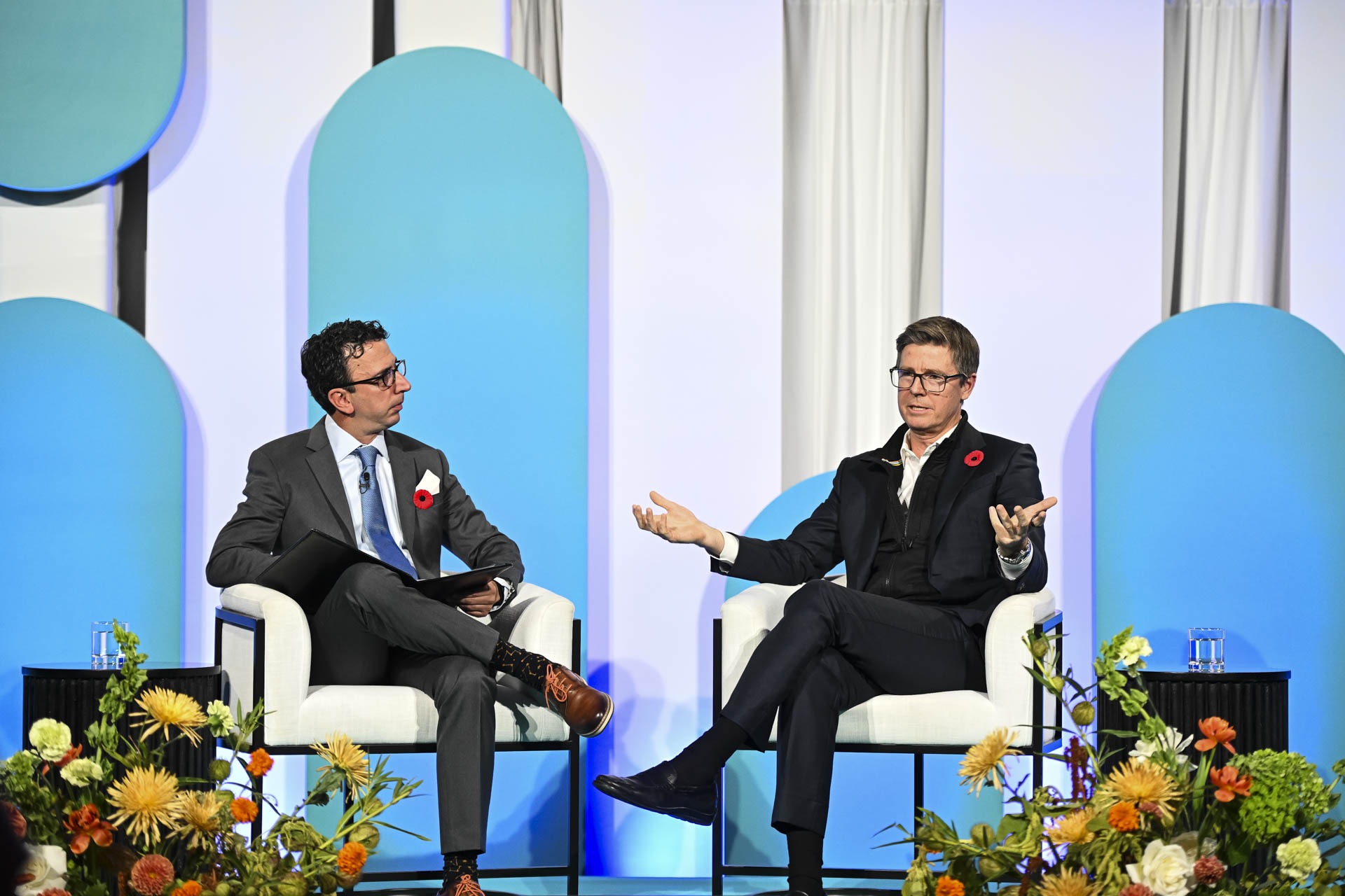 Galen Weston, wearing a dark jacket, black quarterneck and a poppy on his lapel, sits in a white chair with arms slightly raised. The Logic's David Skok looks toward him with one leg resting on the other knee, and a clipboard open. There are flower arrangements at stage level in the foreground.