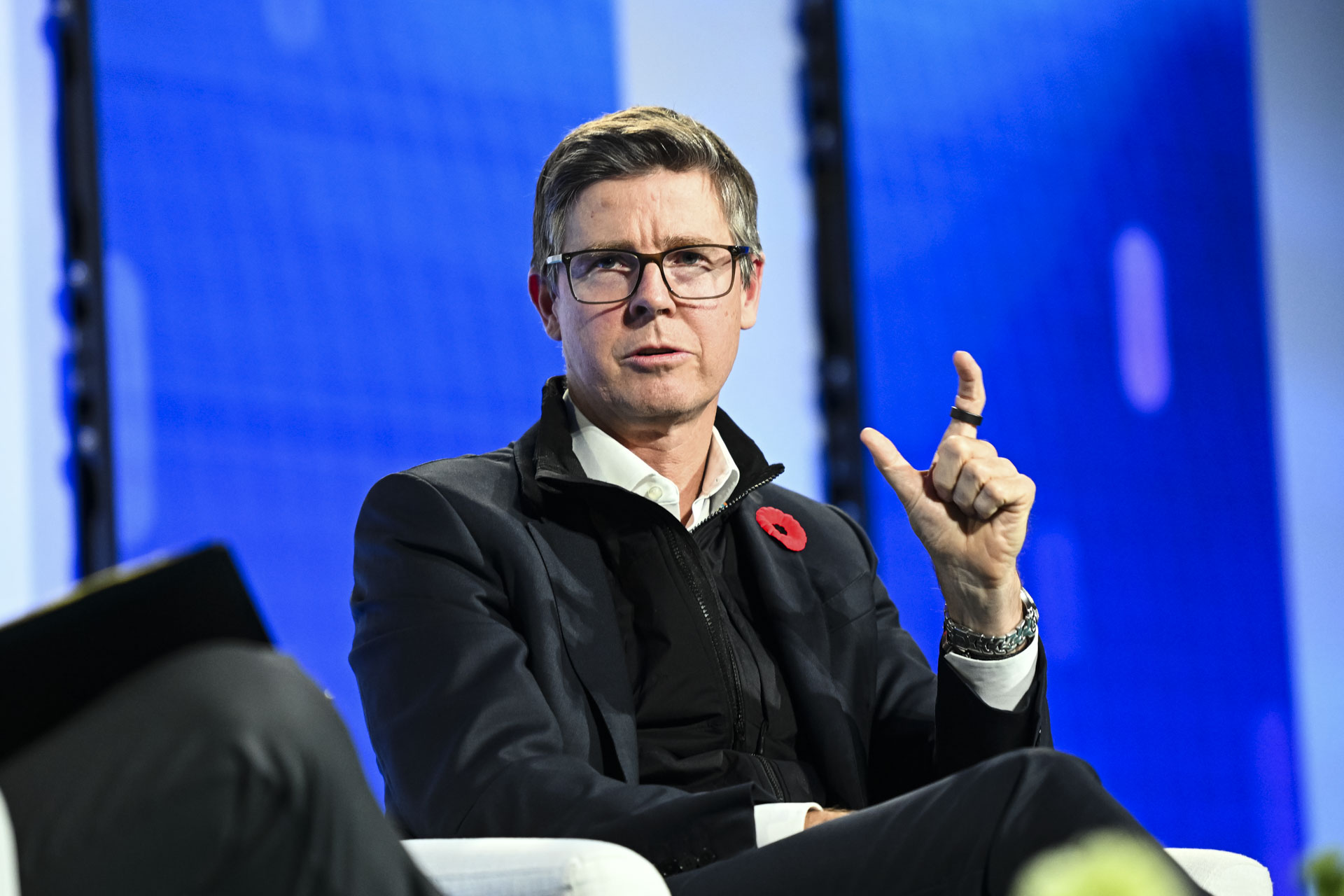 Galen Weston, wearing a dark jacket, black quarterneck and a poppy on his lapel, holds his left hand before him and makes a pincer gesture his thumb and forefinger. There is a black ring visible on his forefinger.