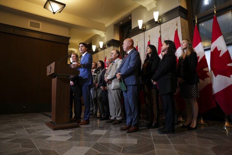 Prime Minister Justin Trudeau and Immigration, Refugees and Citizenship Minister Marc Miller standing with fellow members of Parliament at a podium in a formal room with Canadian flags in the background.
