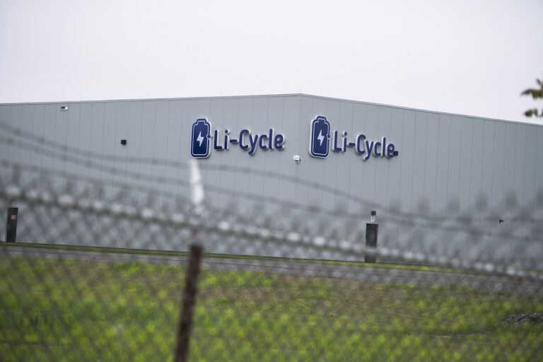 A large building with the "Li-Cycle" logo on its exterior is seen behind a chain-link fence.