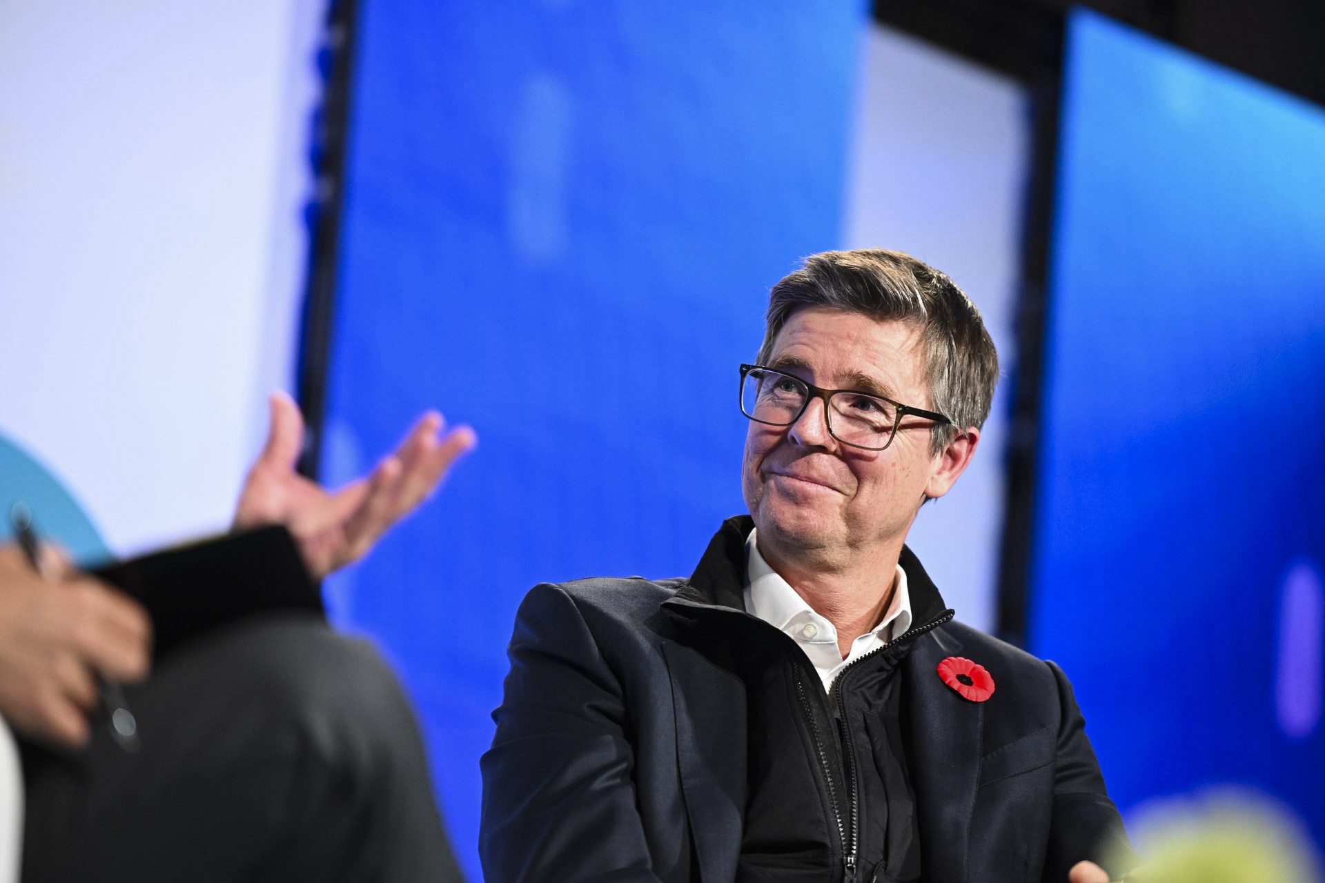 Loblaw chair Galen Weston wearing glasses and a poppy pin, listening attentively during a conversation against a blue backdrop.