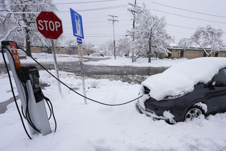 An electric Nissan Leaf is plugged into a charging station while blanketed in snow. A Stop sign and electric charging sign are nearby, and the surrounding streets, buildings and trees are covered in snow.