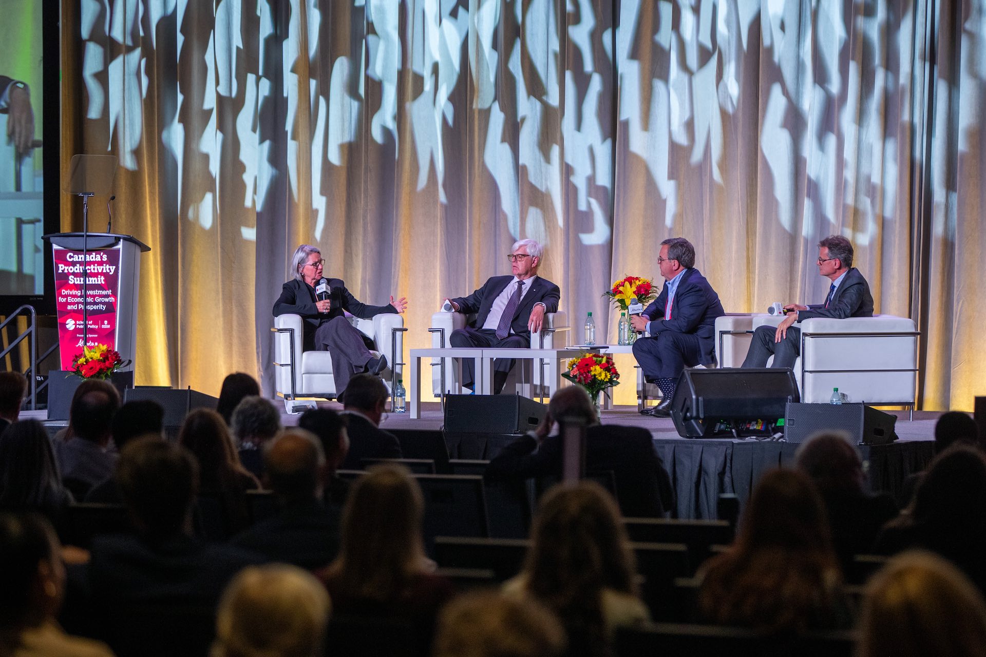 Three male panelists in suits sit onstage while Martha Hall Findlay of the University of Calgary's School of Public Policy asks questions. Findlay, who is holding a microphone, is seated on the far left next to a lectern. The heads of audience members are visible in the foreground.