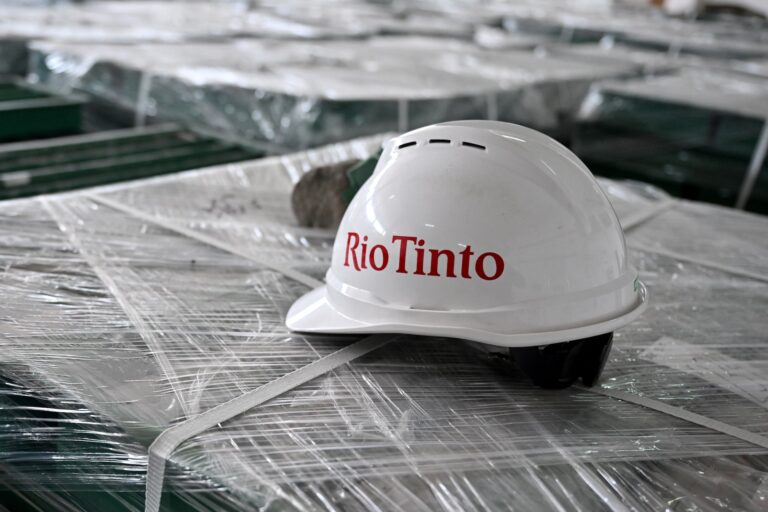 A white hardhat with the Rio Tinto logo in red letters sits on a large tray that is shrink-wrapped with plastic. A grey ore sample is partially visible behind the hardhat.