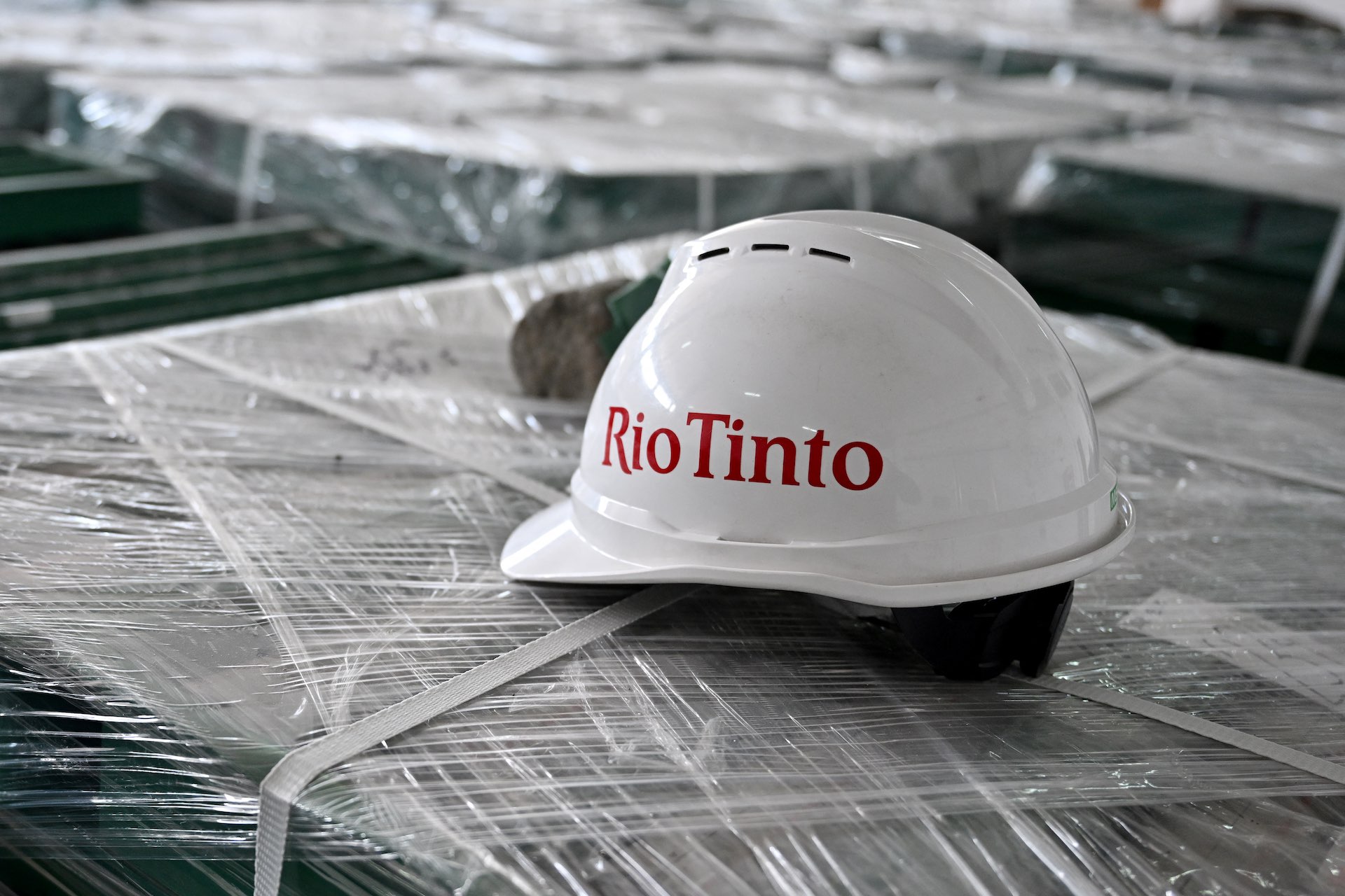 A white hardhat with the Rio Tinto logo in red letters sits on a large tray that is shrink-wrapped with plastic. A grey ore sample is partially visible behind the hardhat.