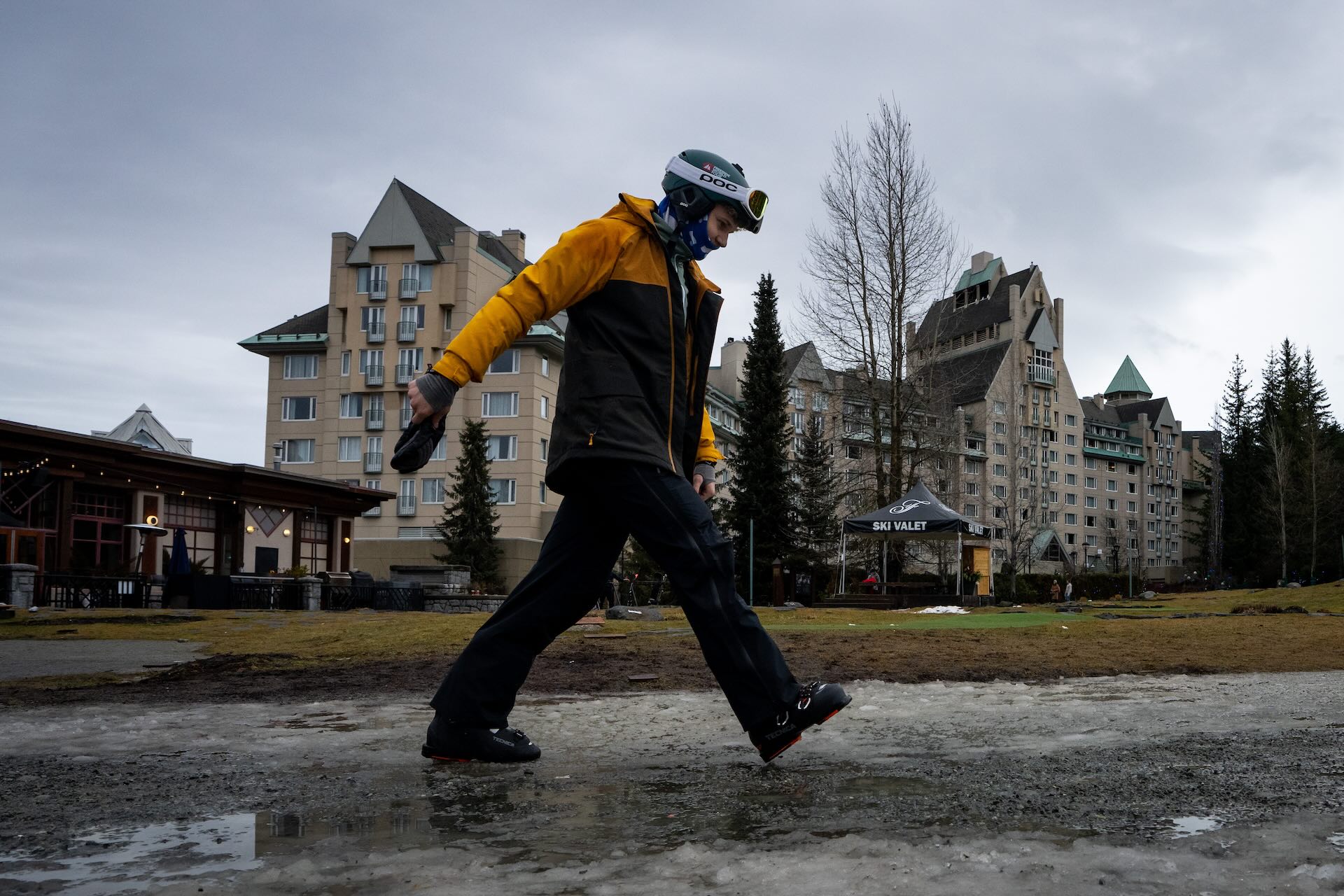 A person in a yellow and black jacket walking on icy ground, holding a phone. There are buildings and trees in the background under a cloudy sky.