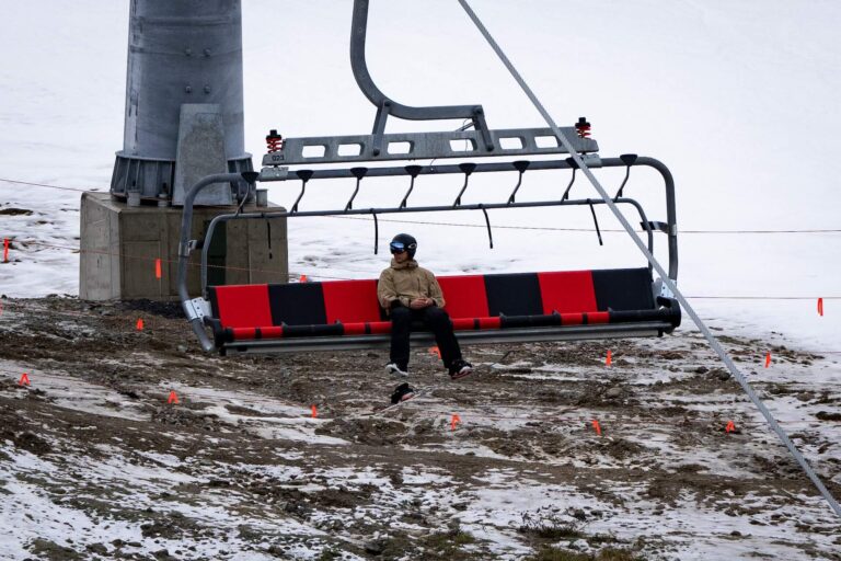 Person seated on a stationary ski lift chair above a rocky, snow-dusted terrain.