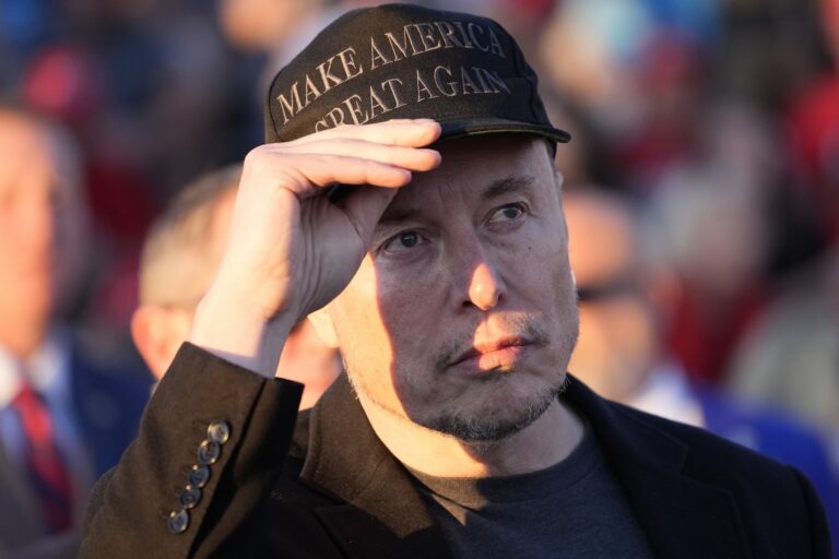 Tesla and X CEO Elon Musk wearing a "Make America Great Again" hat and a black jacket adjusts the hat's brim in a crowd.