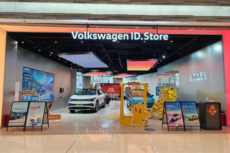 A shot of a storefront inside a mall with three Volkswagen floor-model cars inside. A sign above the store reads "Volkswagen ID.Store." There are Pokemon-themed cutouts and stuffed animals in the foreground—part of an apparent promotion.