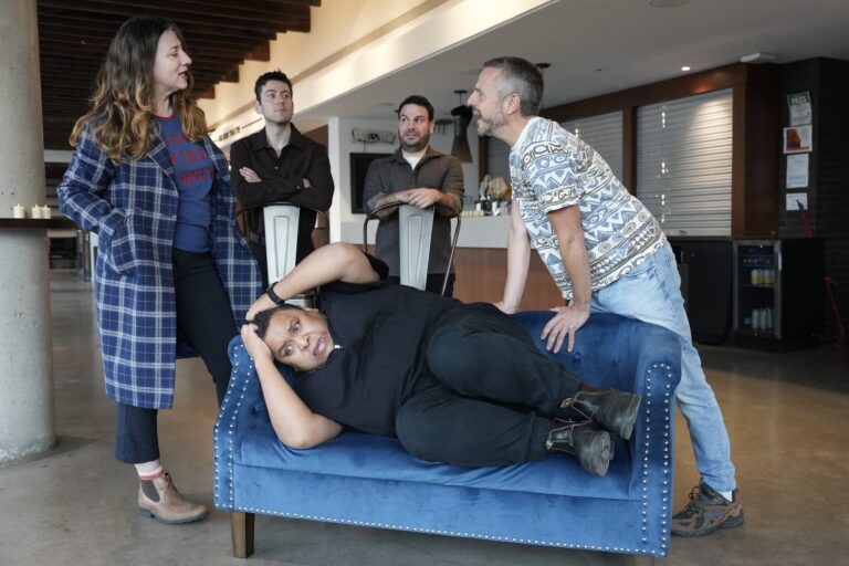 Five people in casual clothes interacting around a blue couch in a spacious room with a high ceiling. One person is reclining on the couch, while the others stand or lean nearby.