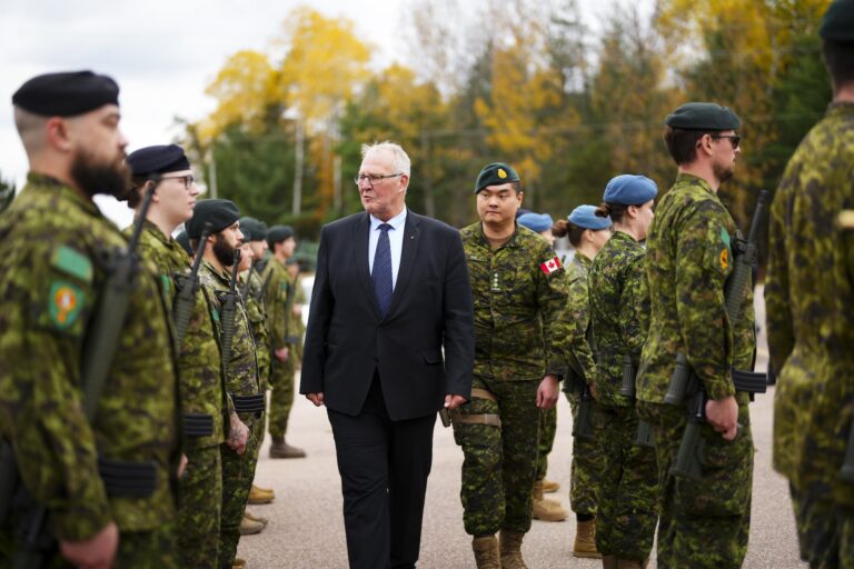 Bill Blair, wearing a dark suit with a blue tie, walks between rows of troops who are standing at attention. Blair is accompanied by a soldier wearing a green beret whose Canadian-flag shoulder badge stands out against his battle fatigues.