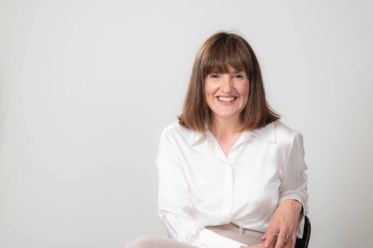 A portrait-style photo of Christine Bellamy, who is seated on a chair with one knee over the other. She is wearing a white blouse, has shoulder-length auburn hair and is smiling toward the camera.
