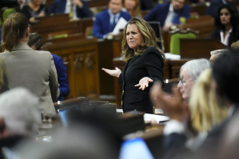 A shot taken from the backbenches of the House of Commons showing Chrystia Freeland theatrically raising both hands and furrowing her brow in puzzlement. She is wearing a black suit jacket with shiny, gold-coloured buttons.