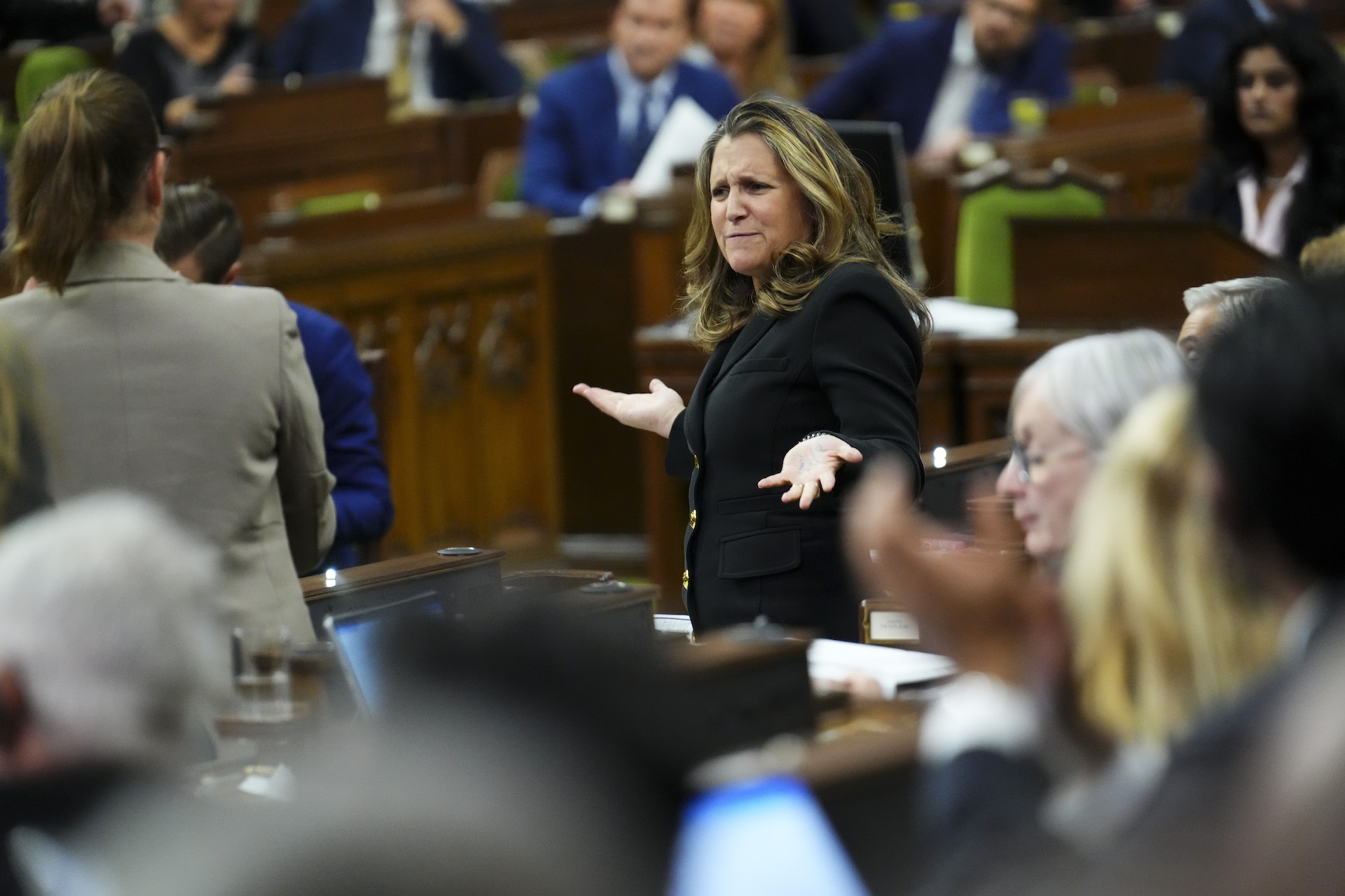 A shot taken from the backbenches of the House of Commons showing Chrystia Freeland theatrically raising both hands and furrowing her brow in puzzlement. She is wearing a black suit jacket with shiny, gold-coloured buttons.