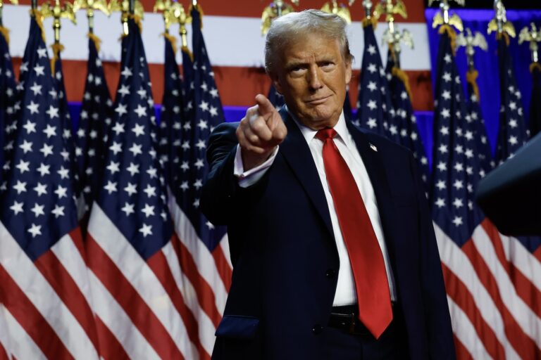 Donald Trump in a suit with a red tie stands in front of a row of American flags and points in the distance