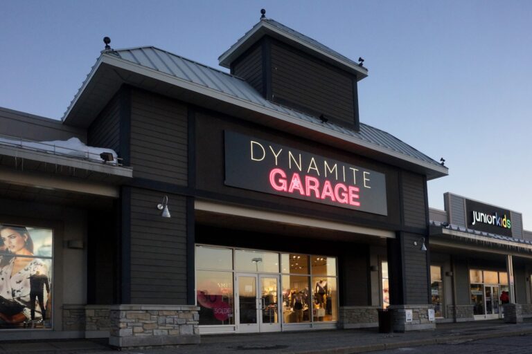 A white Dynamite logo is seen above a Garage store logo, lit in pink. The sign hangs above the door of grey exterior storefront.