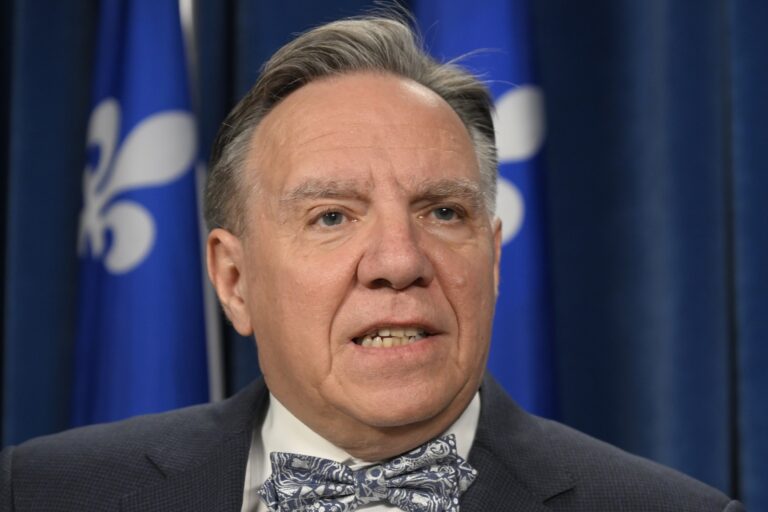 Quebec Premier Francois Legault wearing a suit and bow tie, with the Quebec flag seen behind him.