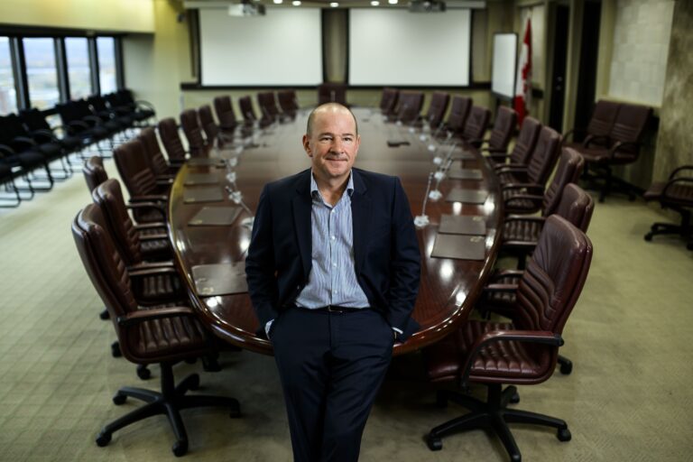 Matthew Boswell leans on the end of a long, oval table that is surrounded by two dozen brown leather chairs in a windowed boardroom. There are small microphones on the table at each setting, and two white pulldown screens in the background.