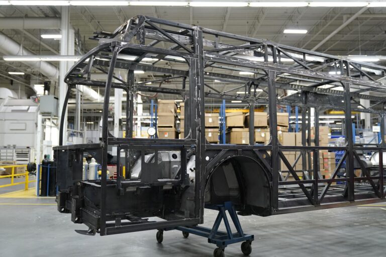The front half of the skeletal steel frame of a bus rests on a dolly, on the floor of a large factory space. Ducts, piping and a large stack of cardboard boxes are visible in the background.