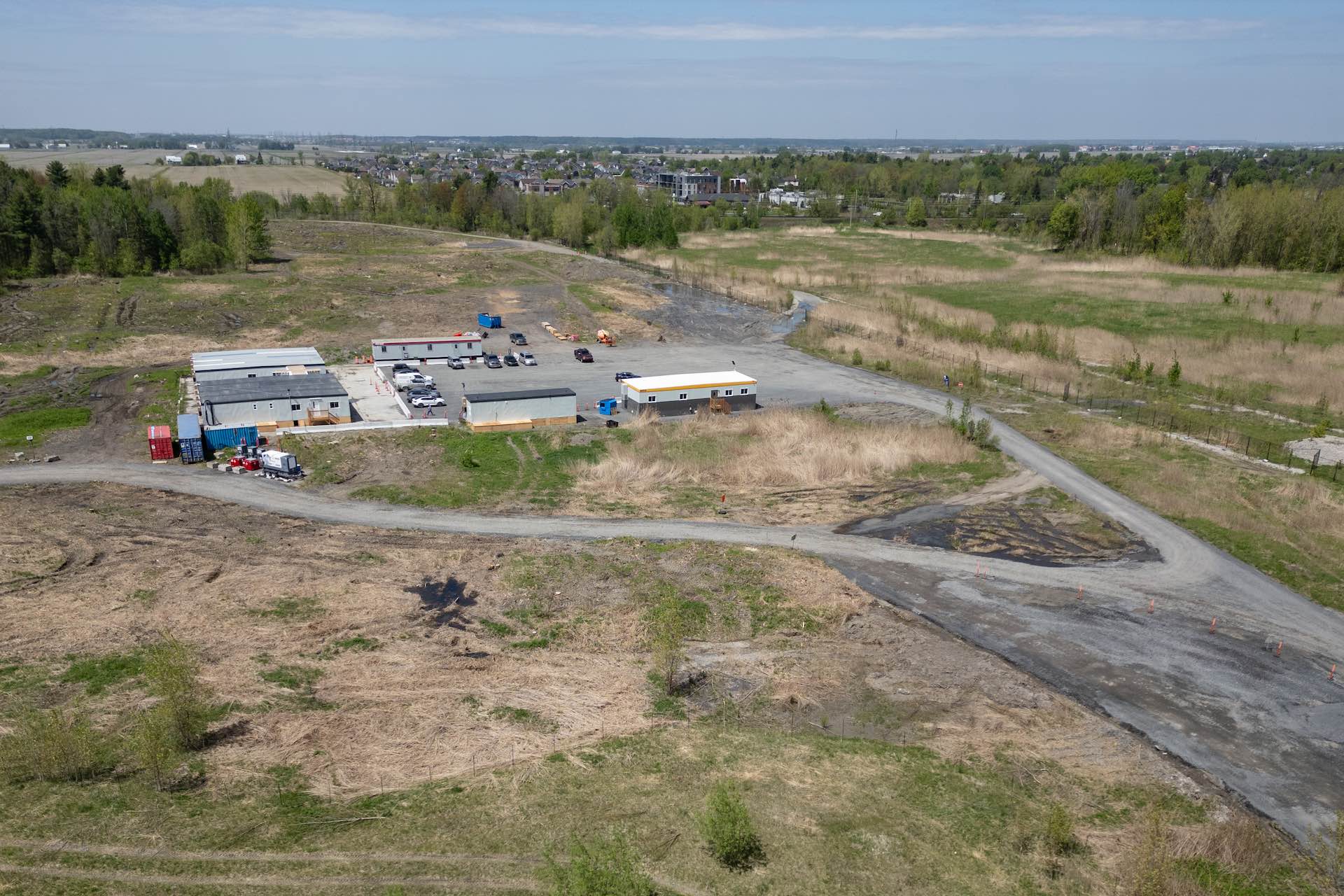 An aerial view of a field with access roads crossing it, and a cluster of office trailers near the centre. The site is banked by low forest and there are buildings in the distance.