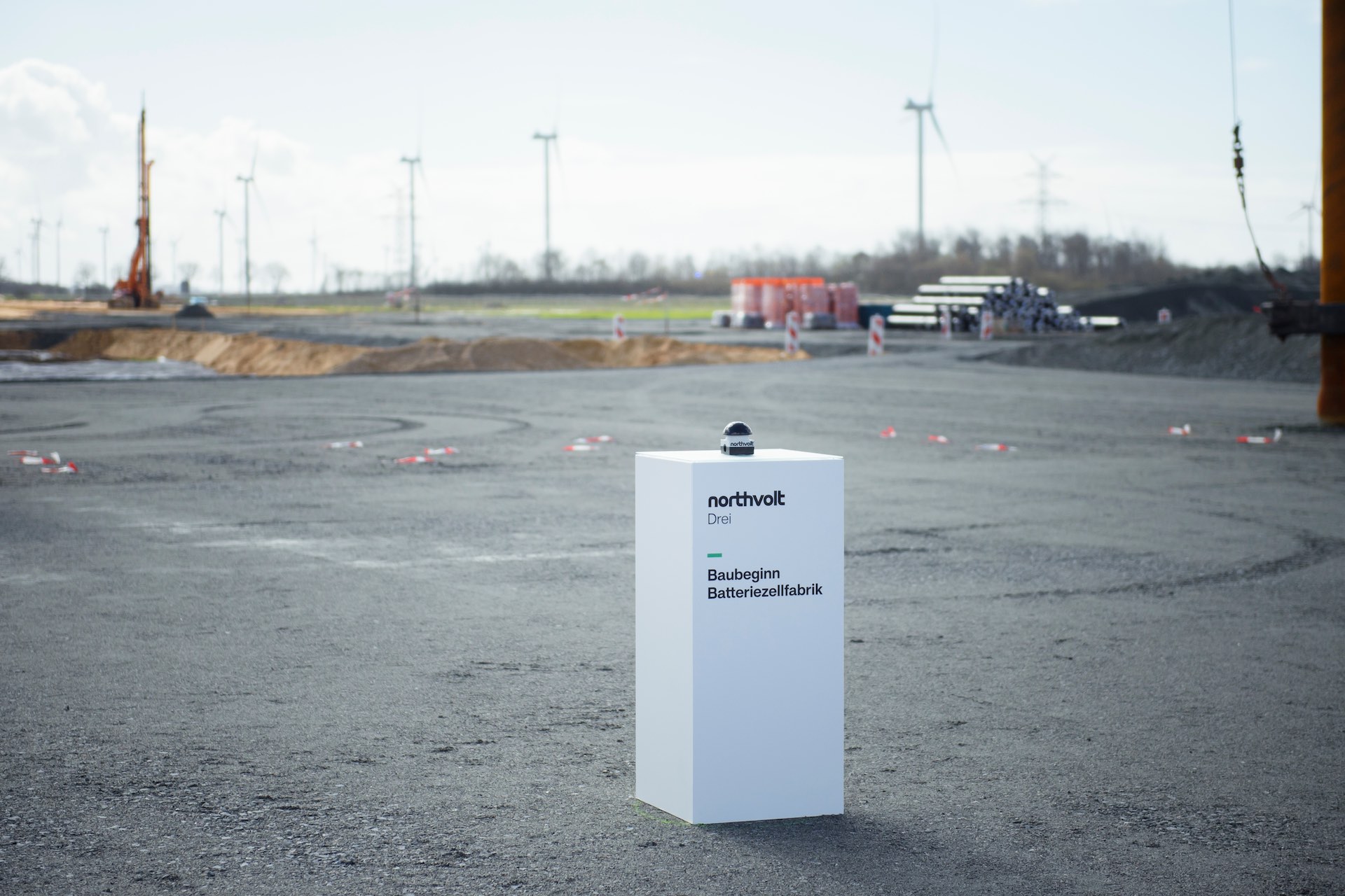 A white podium bearing a black button the size of a half-grapefruit sits unattended in the middle of a vast construction site. A logo on the front of the podium reads "Northvolt Drei" (Northvolt Three).