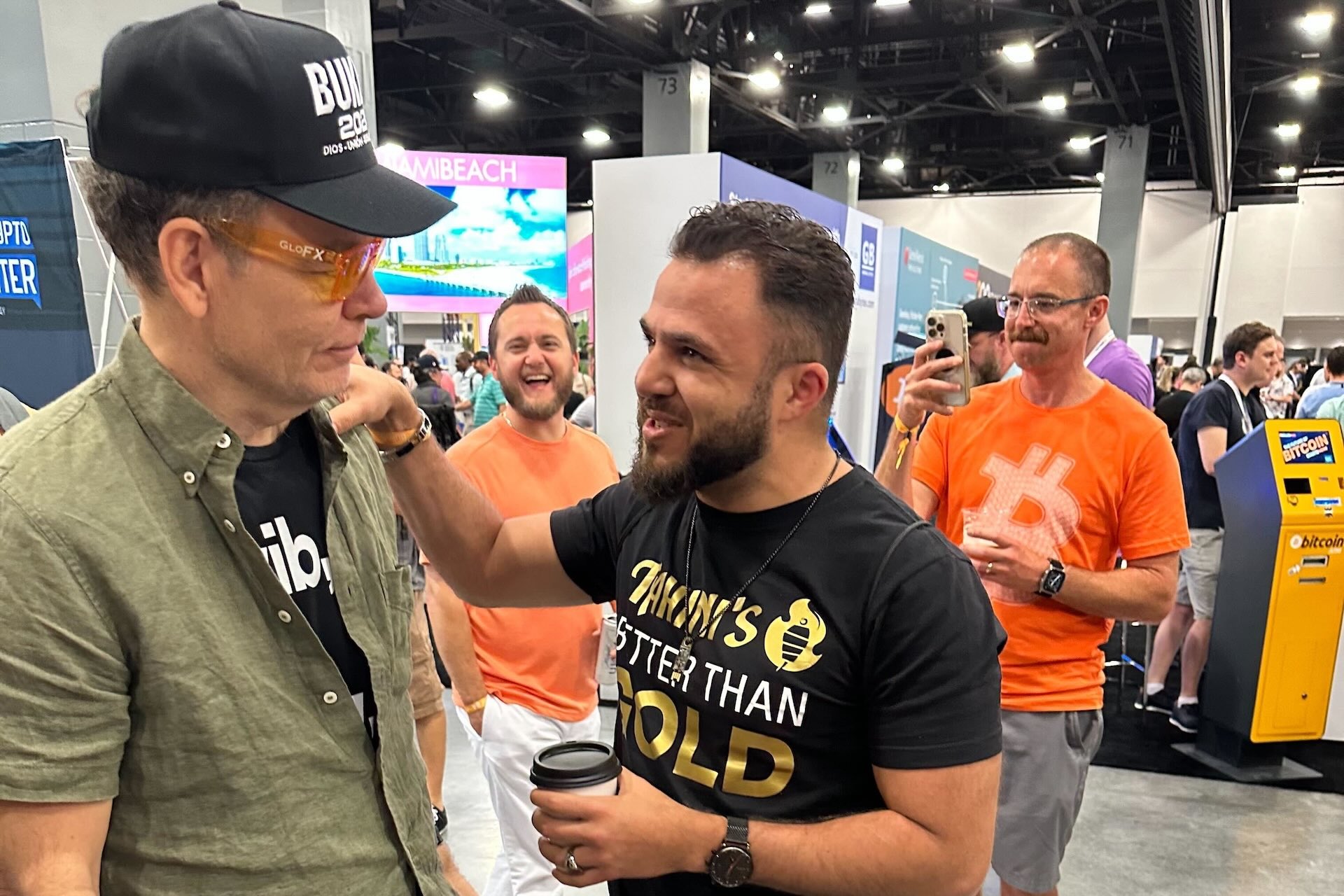 A group of people at a convention center, with two men talking closely in the foreground while others wearing orange Bitcoin T-shirts watch and take photos.