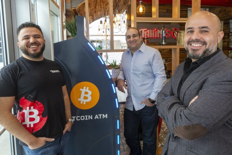 Alt: Tahini’s restaurant co-owners Aly Hamam, Ahmed Dessouki and Omar Hamam stand next to a Bitcoin ATM inside a restaurant.