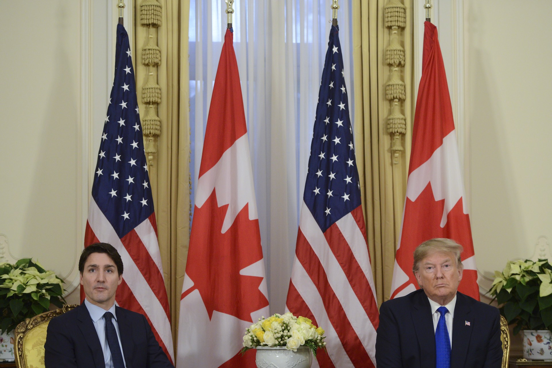 A shot of Trump and Trudeau seated several feet apart with a flower arrangement between them. There are gold-hued curtains and U.S. and Canadian flags behind them.