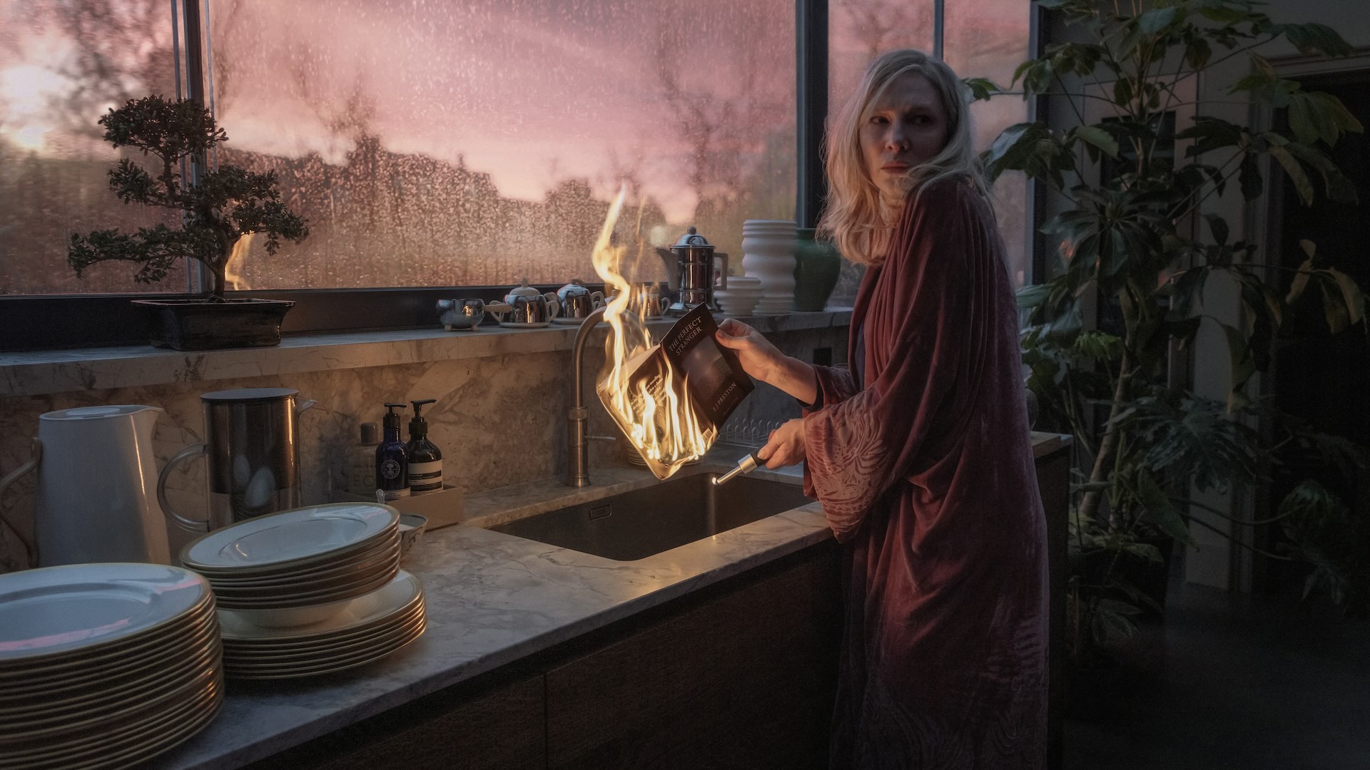 A woman in a purple robe stands over a kitchen sink while she holding a book she appears to have lit on fire, looking over her shoulder while the flames burn