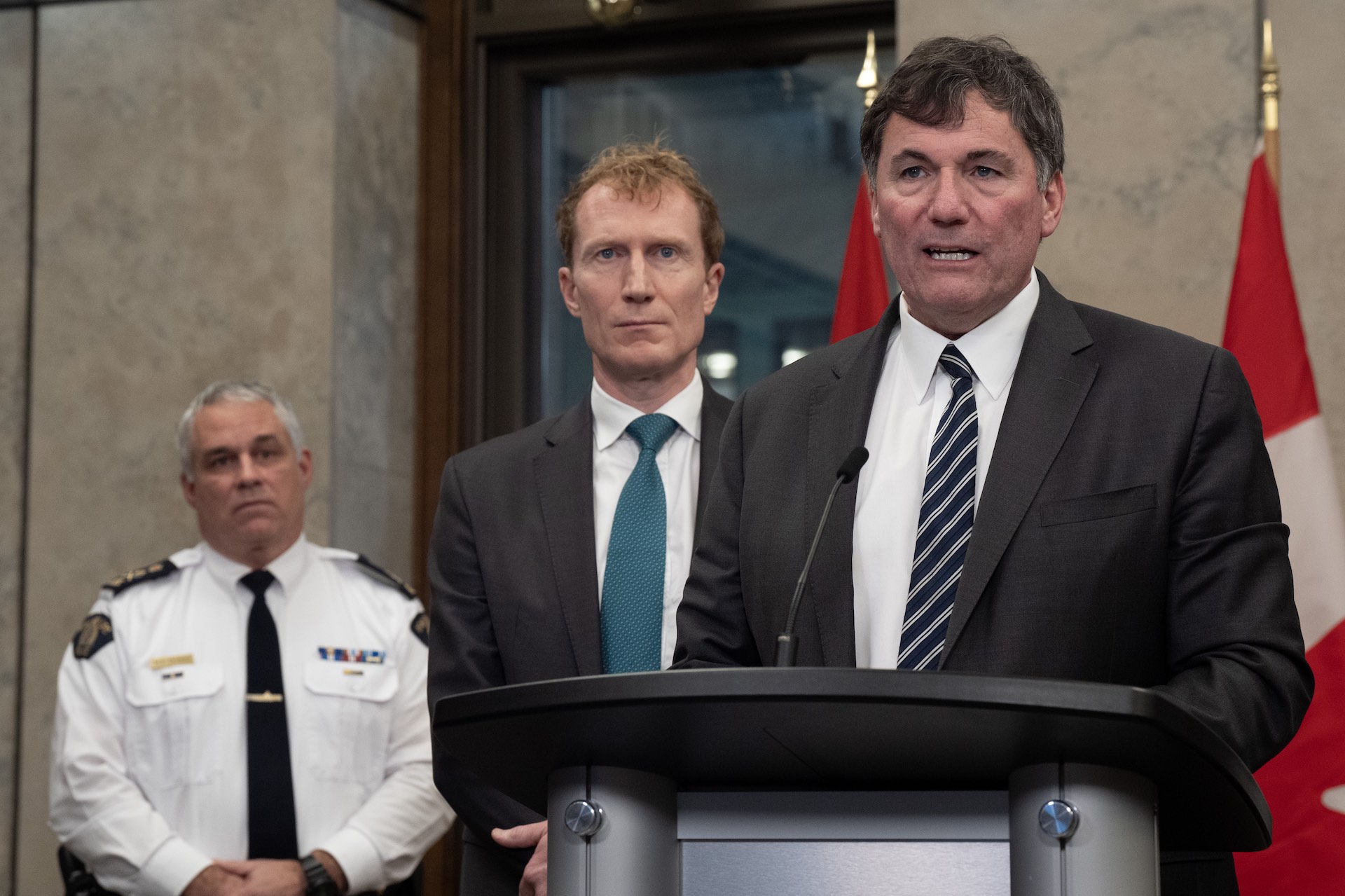 LeBlanc in a dark grey suit and striped tie at a podium; Miller is standing to his right in a suit and teal-coloured tie; on his far right is Duheme, in his RCMP uniform.