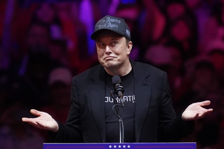 Elon Musk shrugs with hands out to each side while standing behind a podium microphone, wearing a black baseball cap that says "Make America Great Again."