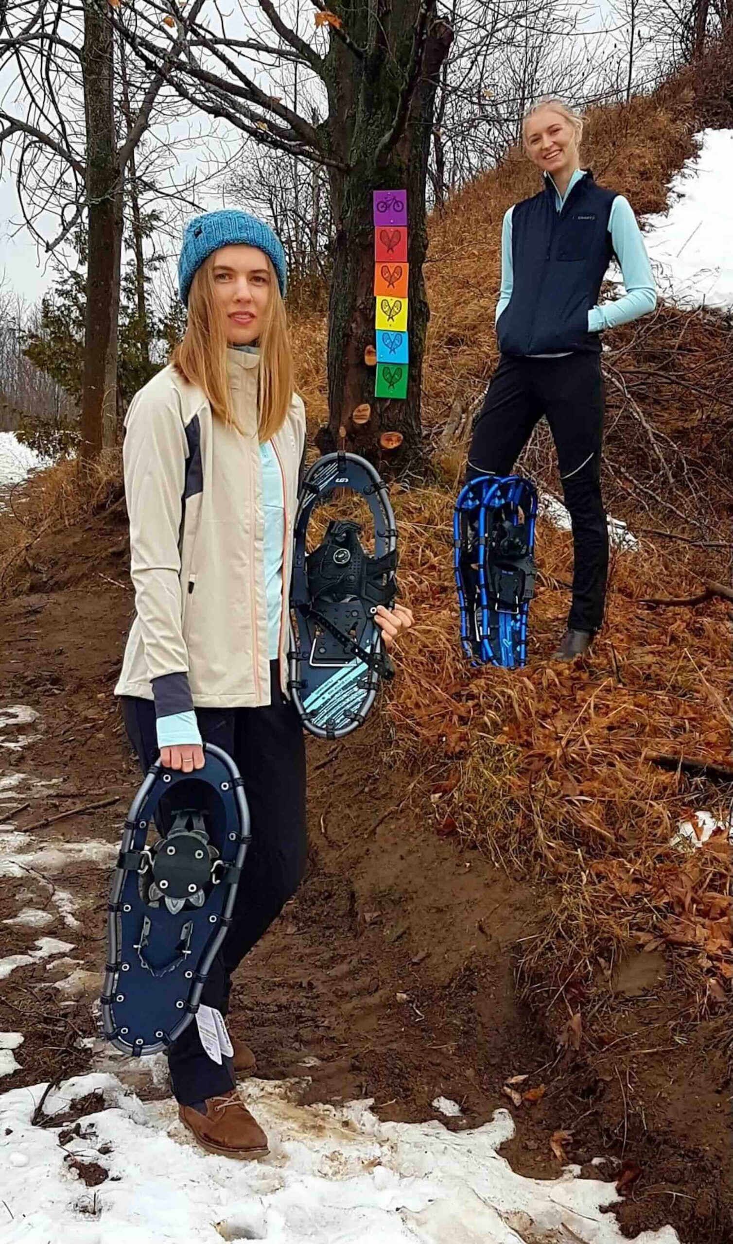 Two women stand on a hill of brown grass, holding snow shoes. Some snow can be seen on the ground.
