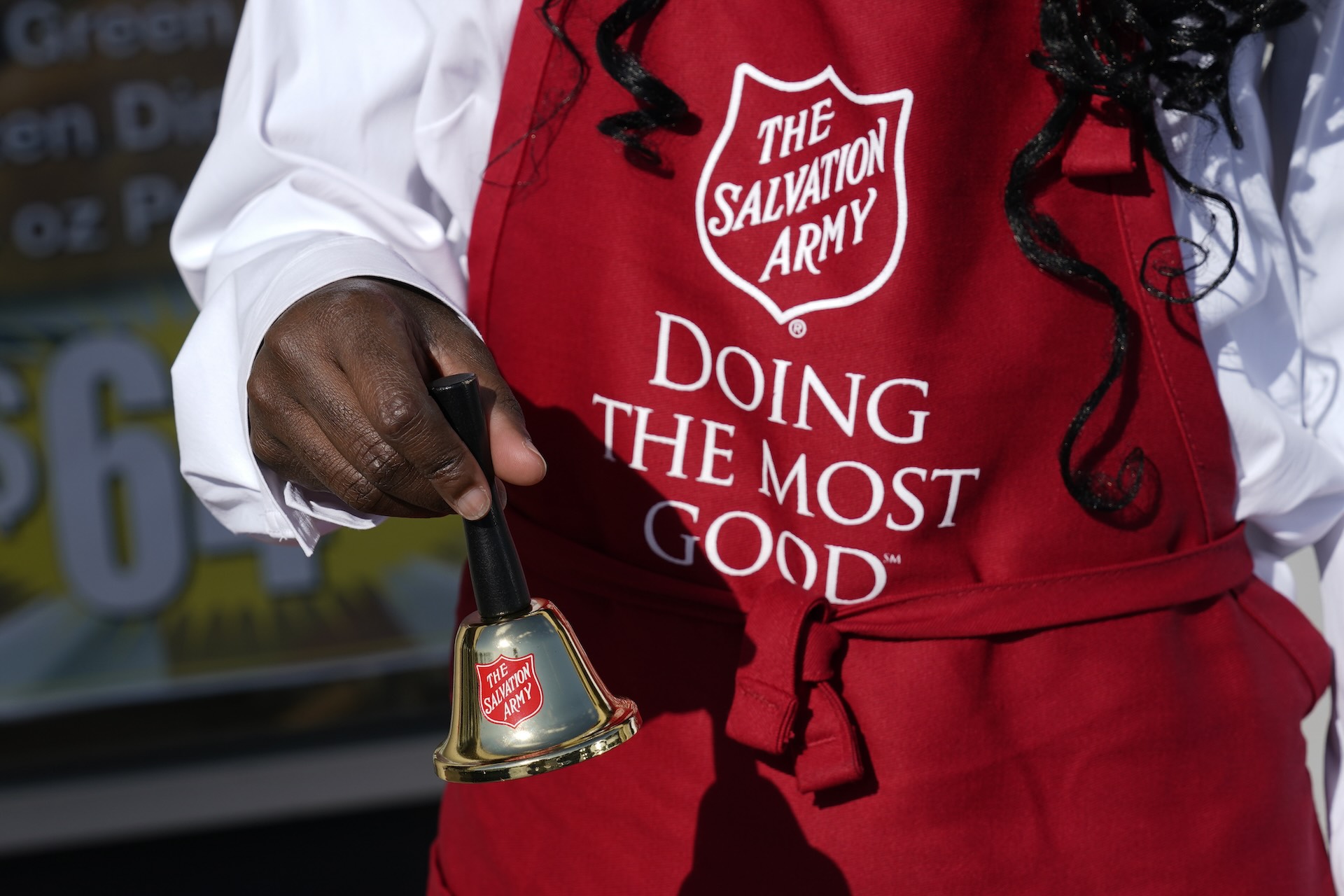 A woman wears a red apron that reads "The Salvation Army" and "Doing The Most Good." The photo is of her hand and the apron, and she is ringing a golden bell