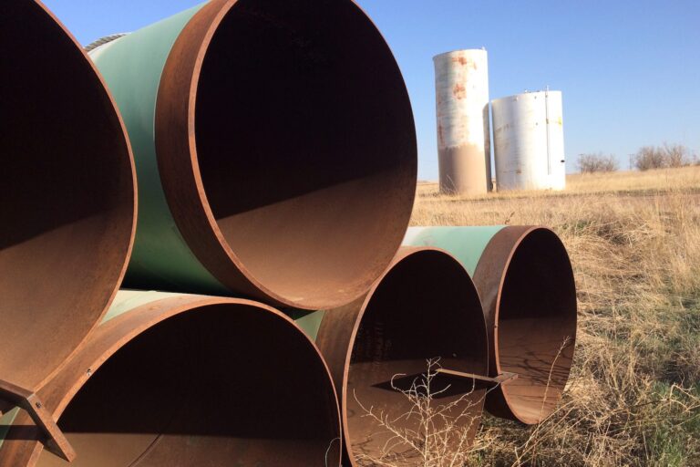 An end-view of segments of steel pipe in a stack with mint-green paint on their outsides and a rusty patina on their insides. There are weeds growing around the pipes and two cylindrical white tanks are standing in the background.