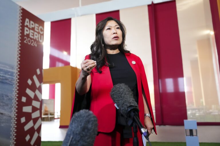 Trade Minister Mary Ng speaks to reporters while wearing a bright red suit. A sign behind her says “APEC Peru 2024.”