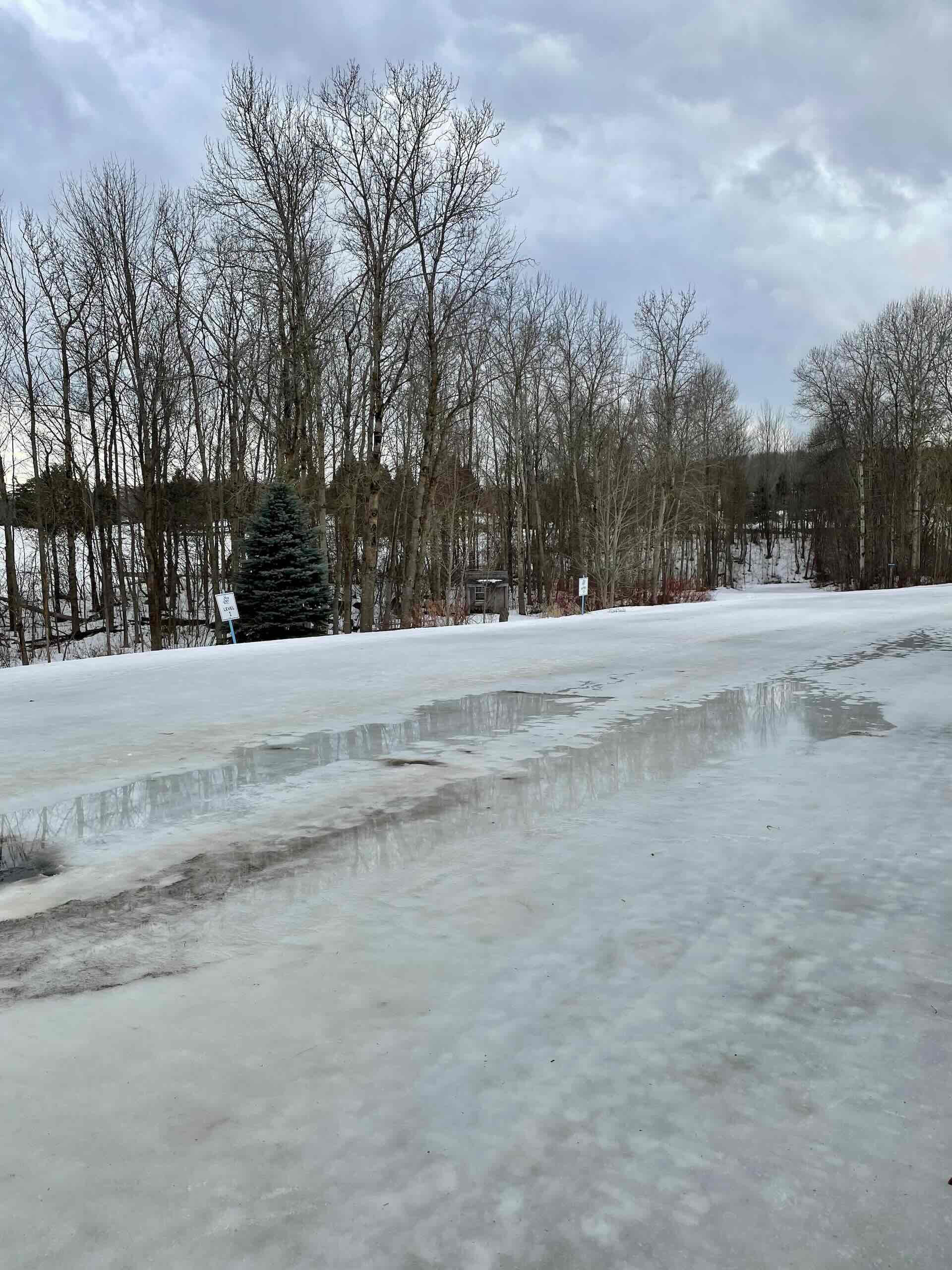 Snow on a cross-country ski trail is melting. Bare trees are seen in the background.