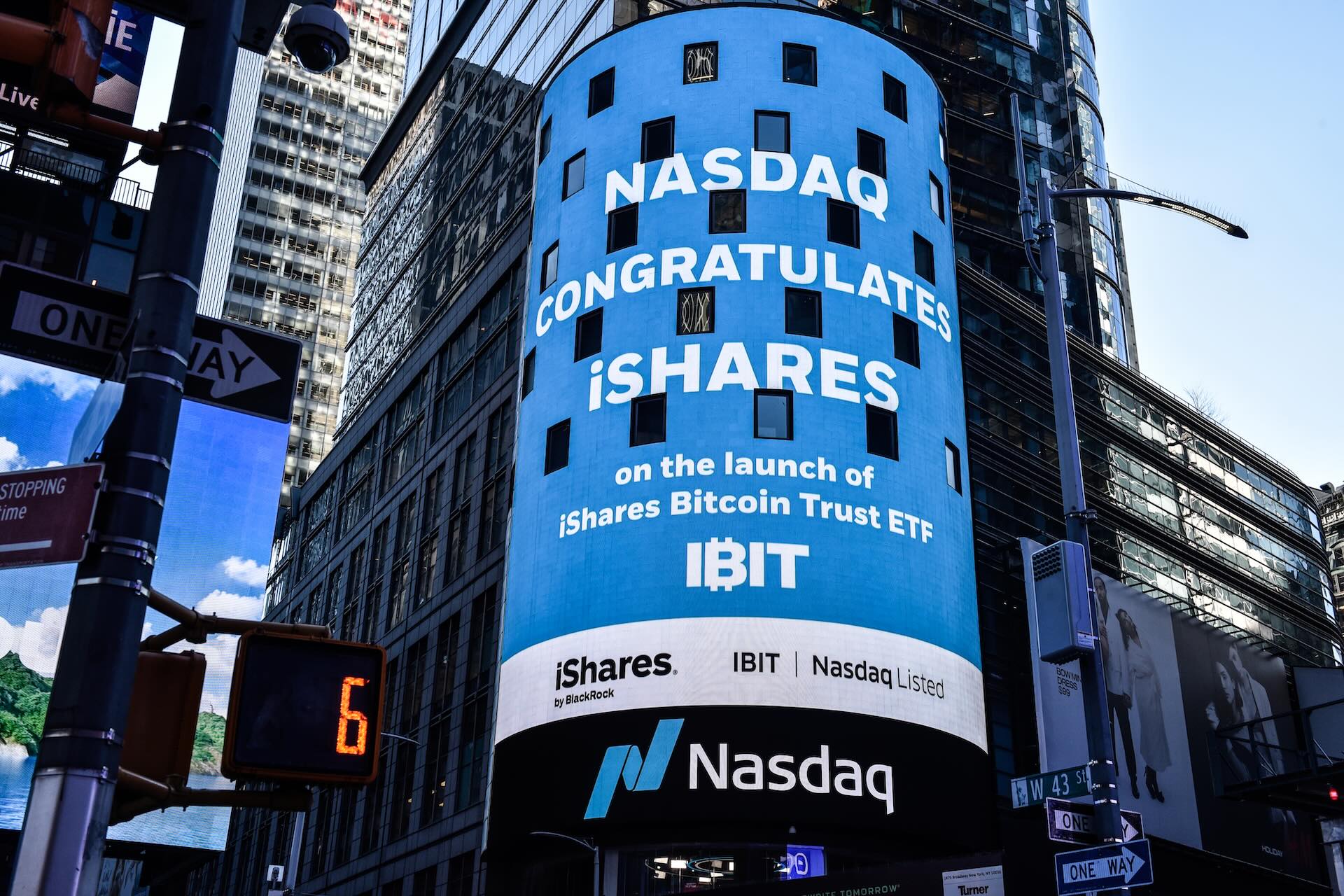 A large blue screen showing the Nasdaq Exchange in Times Square says, “Nasdaq congratulates iShares on the launch of iShares Bitcoin Trust ETF,” in white text. Road signs and skyscrapers are seen around the screen.