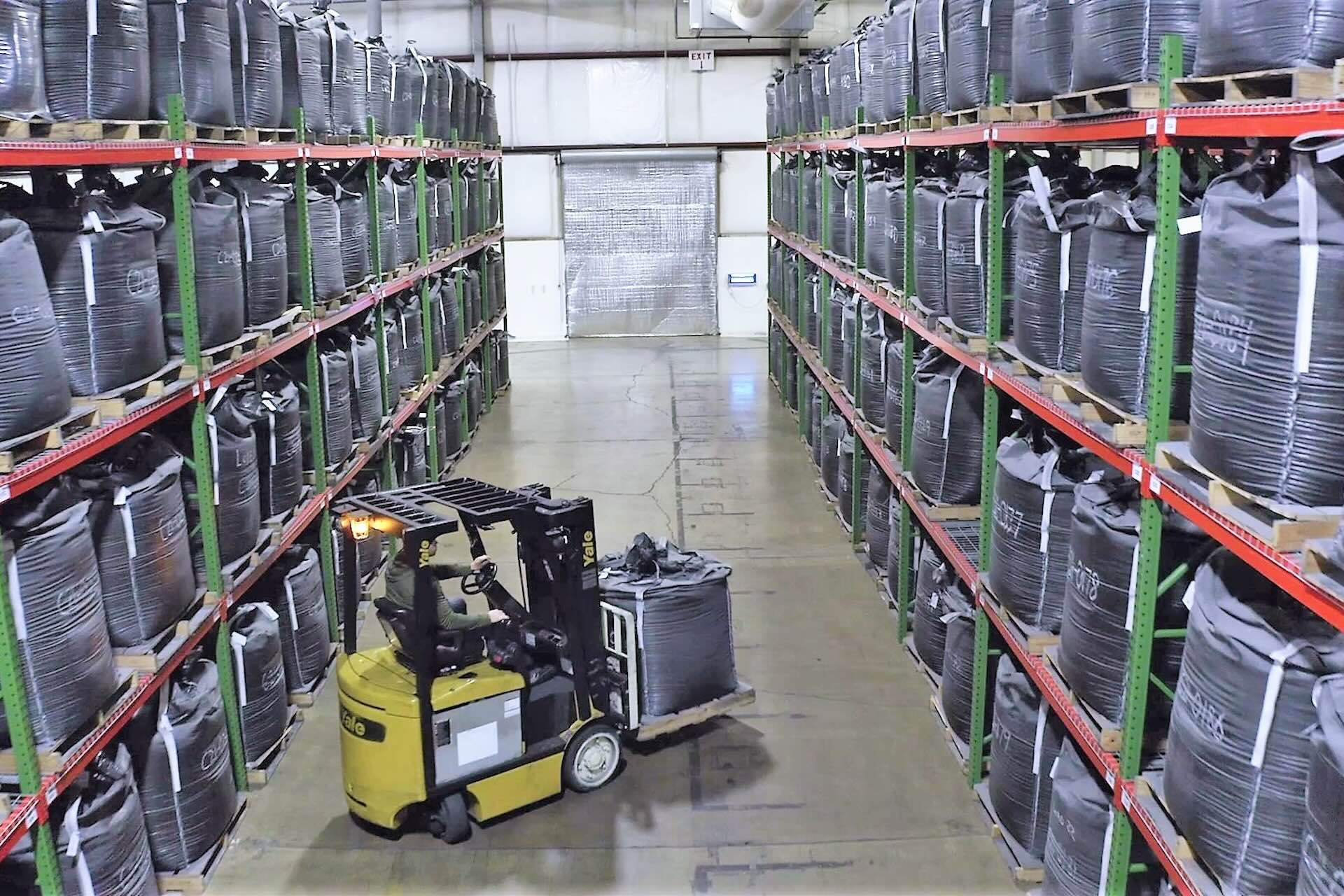 A forklift between two tall shelves with dozens of large black bags of graphite. The cart is transporting a large bag.