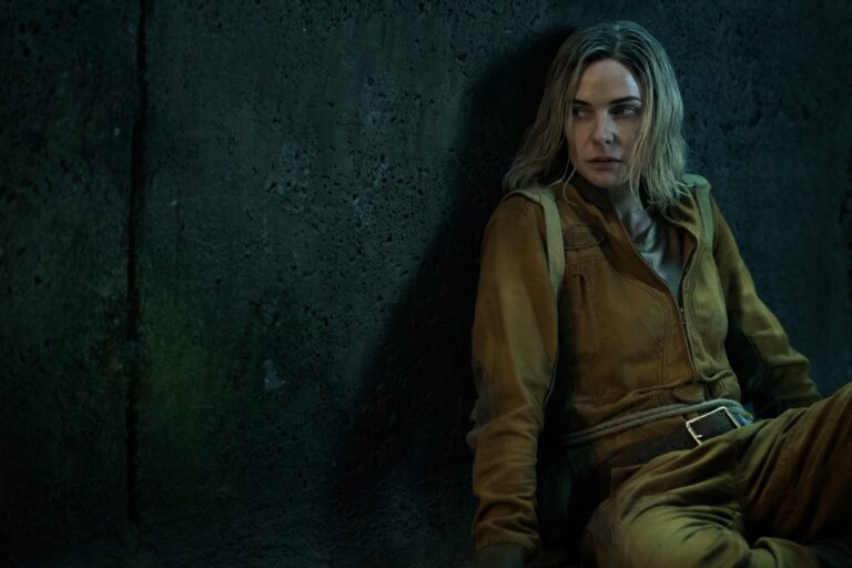 A blonde woman in a dirty yellow jumpsuit leans against an concrete wall, looking off camera warily
