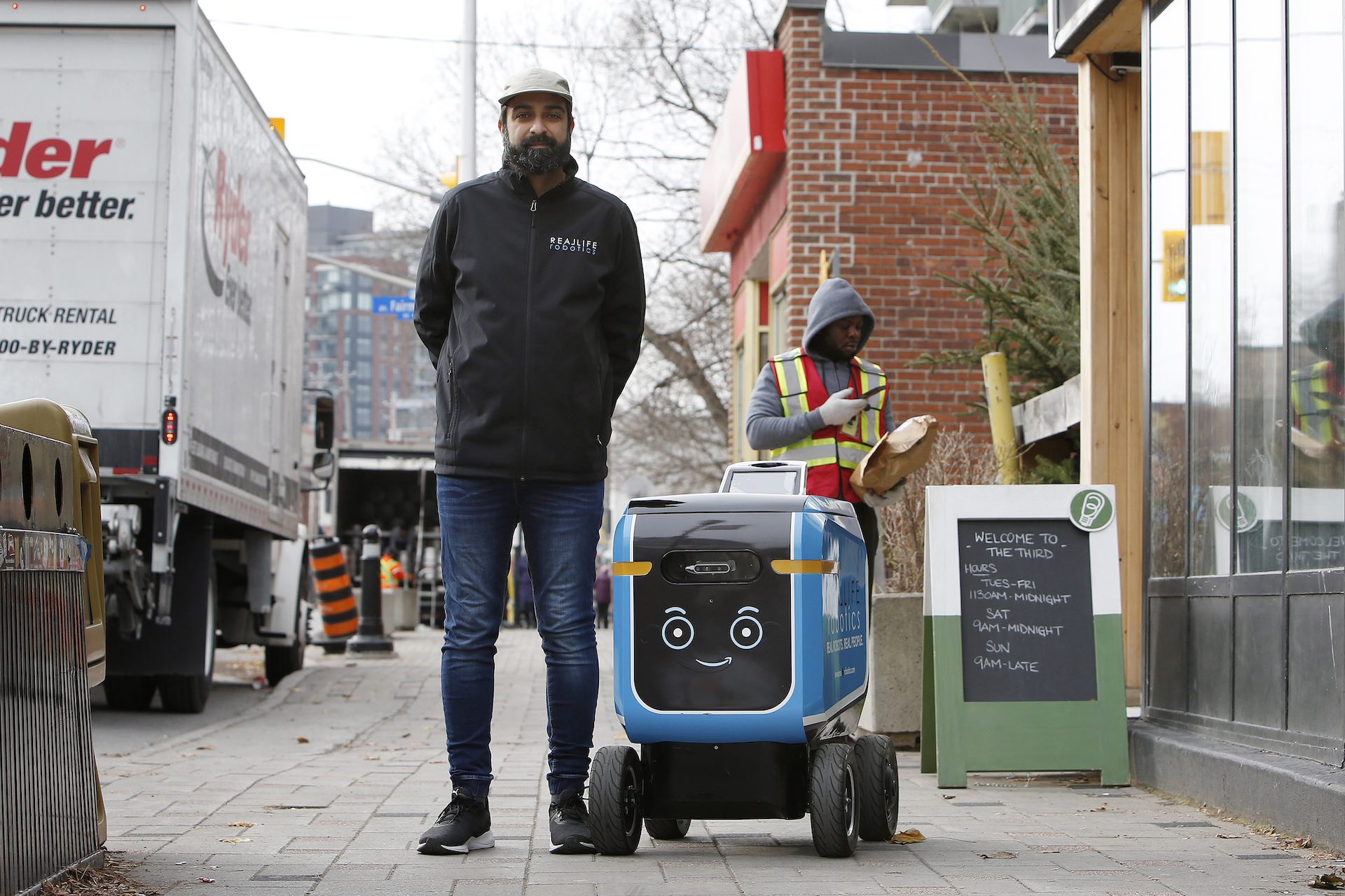 A man with a beard, black jacket and tan-coloured cap stands on a sidewalk beside a robot that looks like a large camp cooler on wheels. In the background is a delivery worker in an orange and yellow reflective vest scanning a parcel code with his phone.