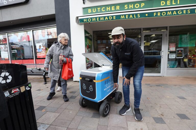 A man with a beard, black jacket and tan-coloured cap stoops slightly beside a delivery robot that looks like a large camp cooler on wheels. They are on a sidewalk in front of a small grocery store. An elderly woman carrying a red shopping bag is peering beneath the open lid of the robot's cargo chamber.