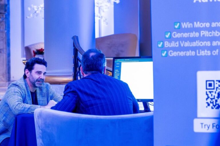 Two men sit across from each other at a table with a computer screen displaying content. A sign nearby lists features like win more and generate lists.