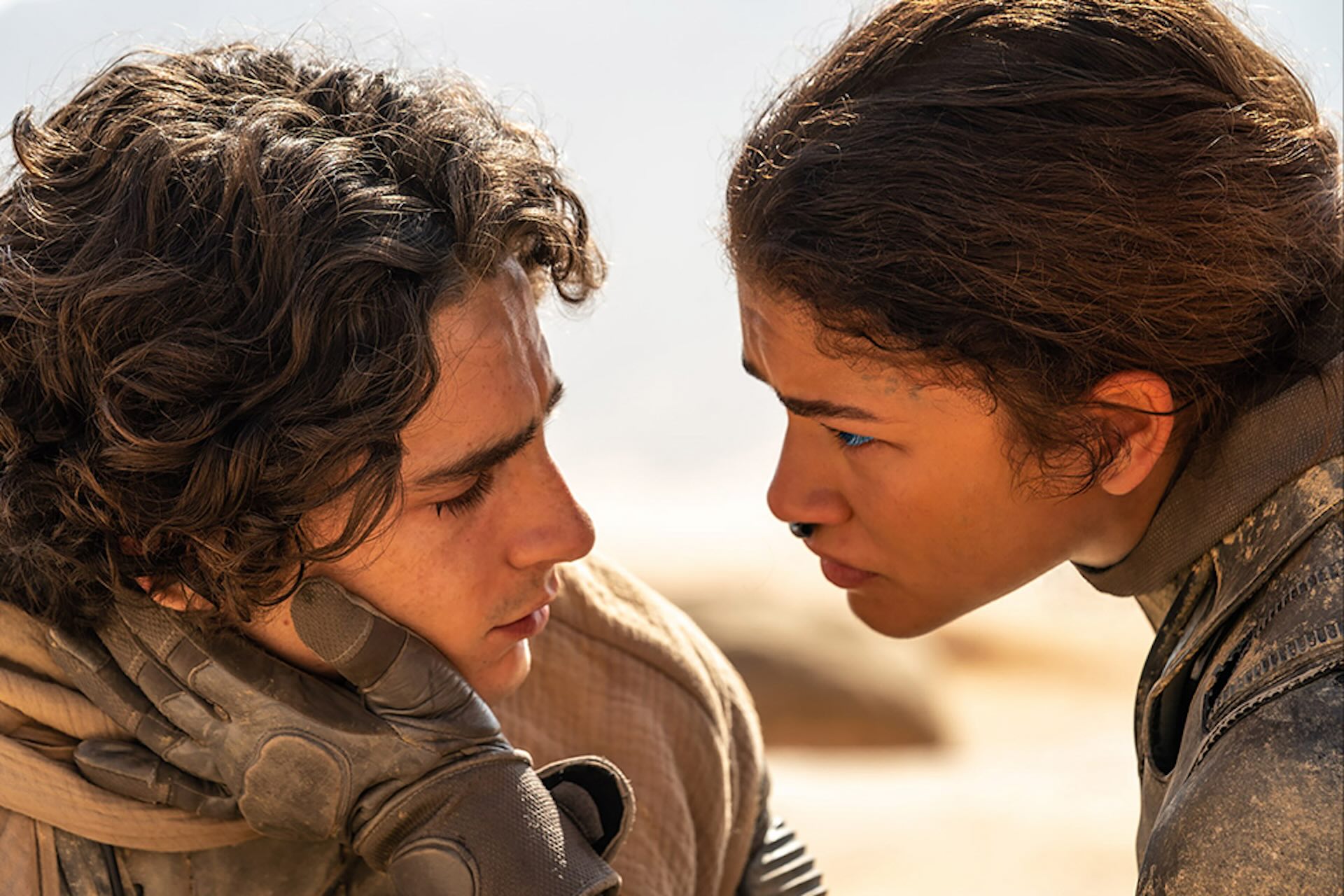 A young woman holds the face of a young man with her gloved hand in a desert.