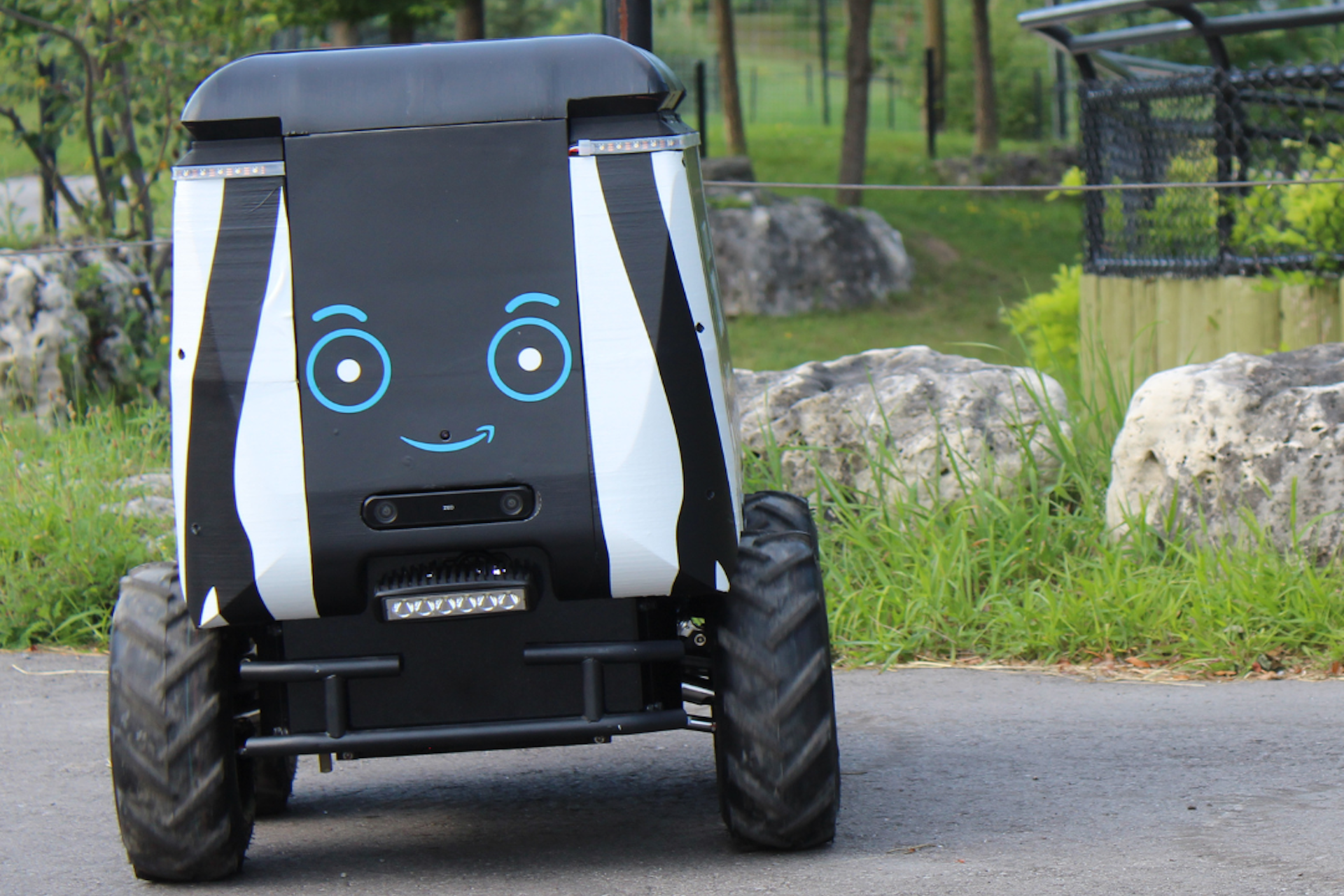 A black-and-white painted robot with wheels and a happy face on its front roll along a walkway in front of an animal enclosure. The robot looks like a large camp cooler on tiny tractor tires. One end of an animal pen is visible in the background.