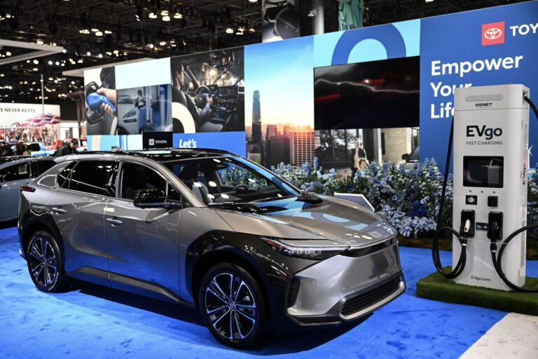 A shining, grey Toyota electric vehicle at an auto show next to a fast-charging station. A billboard sign behind it says "Empower Your Life."