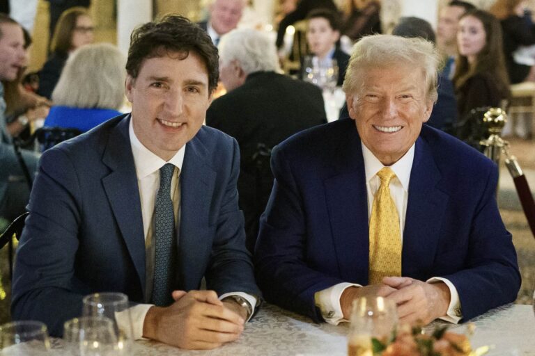 Justin Trudeau and Donald Trump sit side-by-side at a table covered by a white tablecloth, with flowers and drinking glasses in the foreground. Trudeau is wearing a dark suit with a blue tie; Trump a blue jacket and yellow tie. There are people seated at tables in the expansive, ornate dining room in the background.