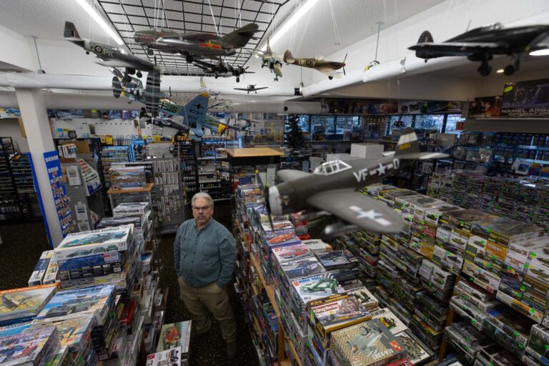 A high-angle photo of a man with grey hair, glasses and goatee standing in a hobby store surrounded by stacks boxes of model cars, planes and ships. There are several assembled models of Second World War-era fighter planes hanging from the ceiling.