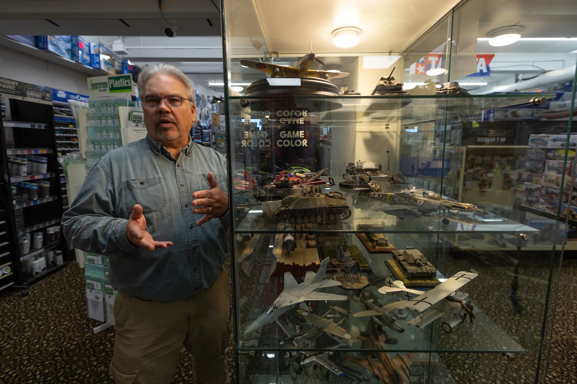 A man with grey hair and a goatee stands by a glass case containing assembled models of tanks and warplanes. He is looking toward the camera and speaking.