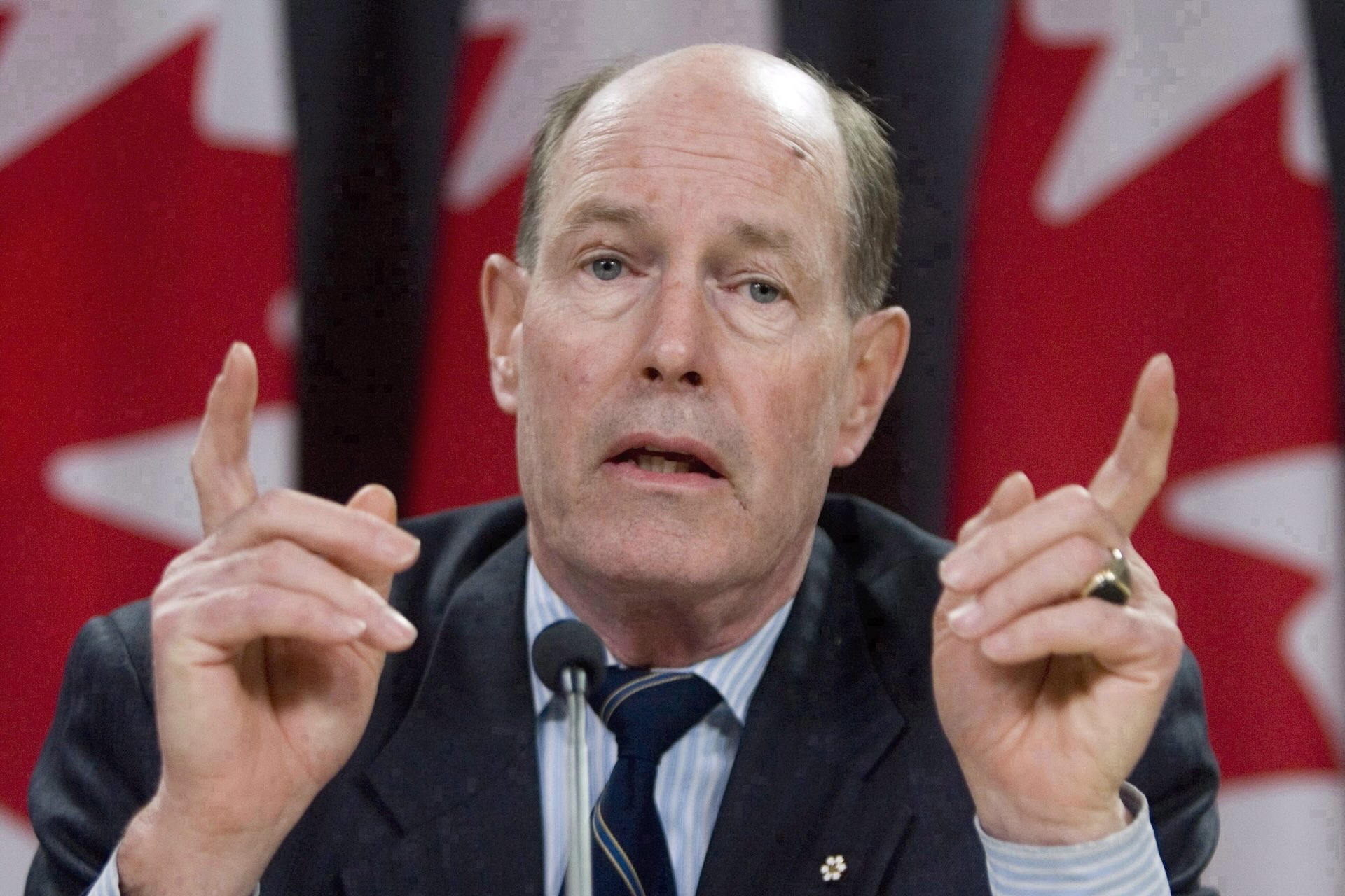 David Dodge, wearing a suit, is sitting and pointing his fingers in the air as he speaks. Canadian flags are in the background.