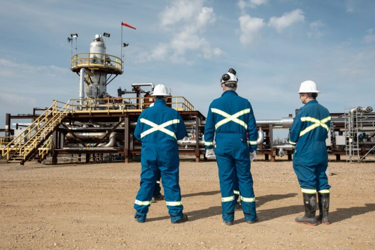A view of oil extraction equipment consisting of pipes, catwalks and cylindrical tanks; there are three company representatives in the foreground wearing white hard hats and blue coveralls with yellow reflective striping.